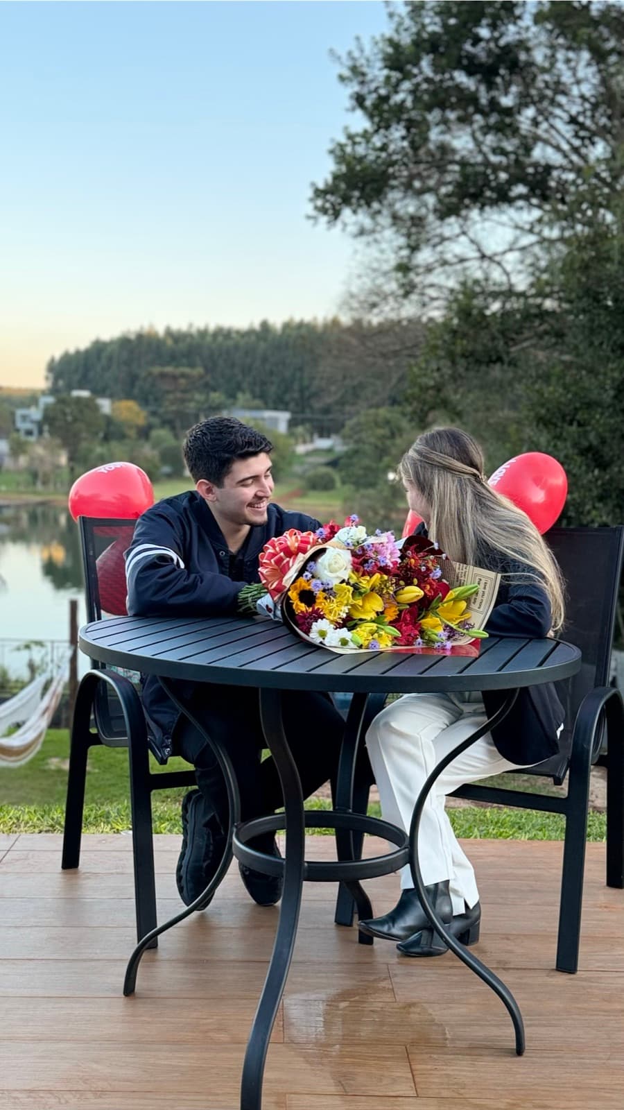 Lívia e Guilherme sentados a uma mesa à beira de um lago no fim da tarde, com um grande buquê de flores e balões