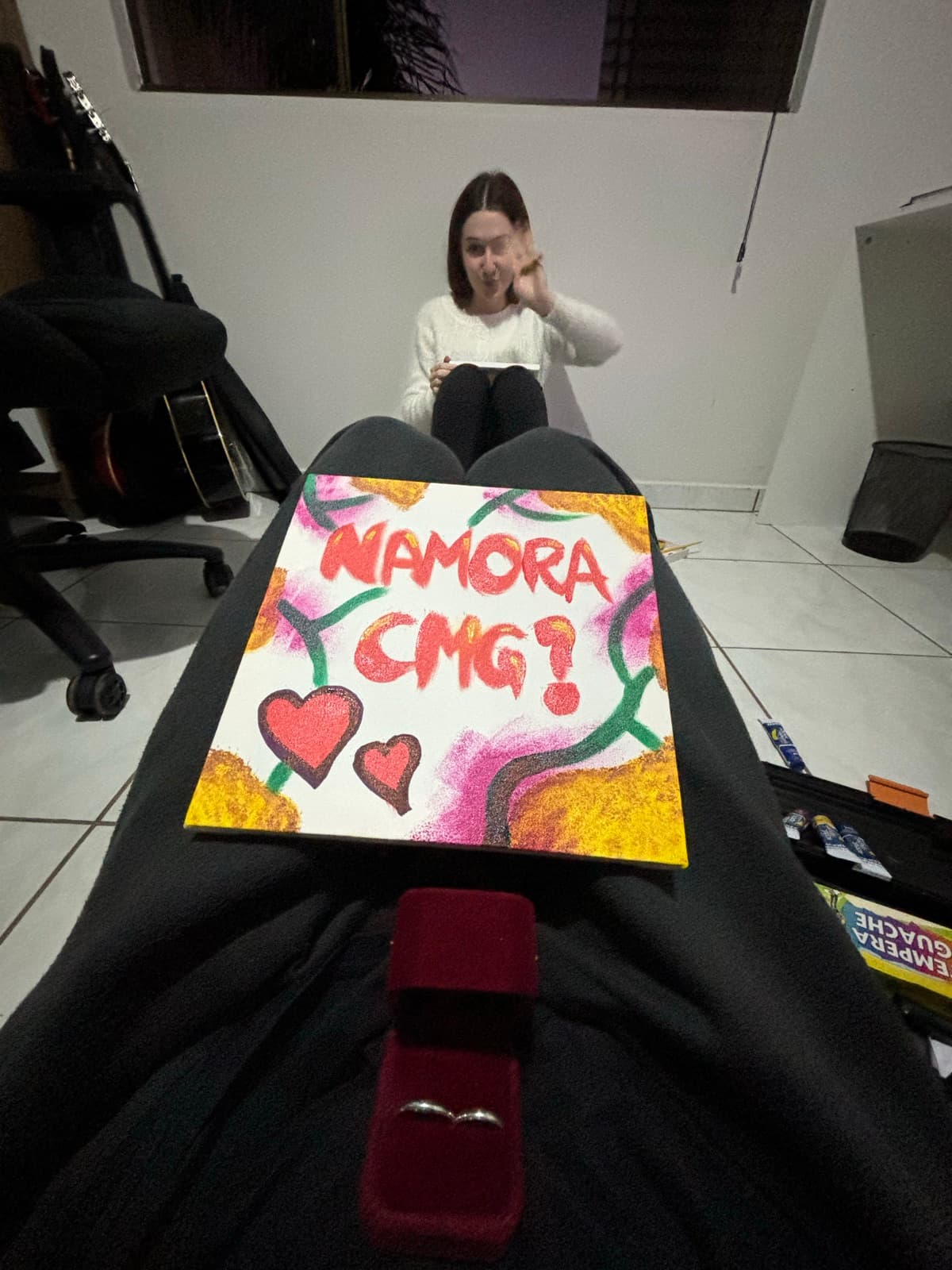 Quadro pintado à mão com a frase “Namora cmg?” e corações, ao lado de uma caixinha de anel vermelha